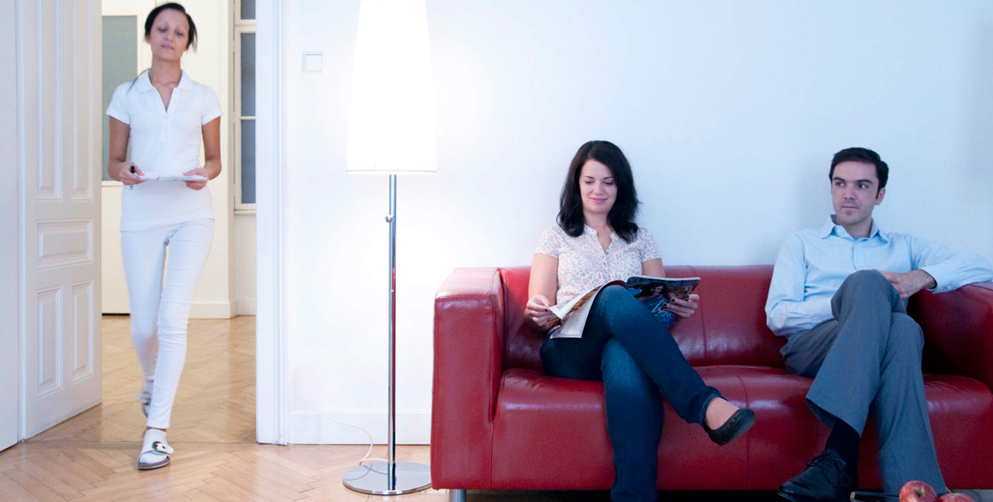Patienten sitzen am Sofa im Warteraum. Assistentin betritt den Raum.